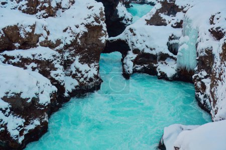 Winterbach mit türkisfarbenem Wasser, das durch schneebedeckte Felsen und unter einer natürlichen Brücke fließt. Ort: Hraunfossar, Island.