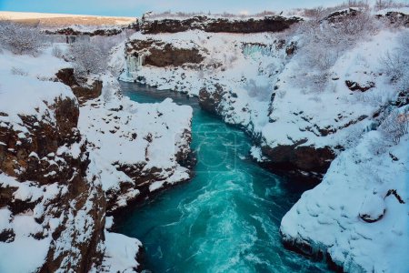 Paysage hivernal d'une rivière gelée avec des berges enneigées sous un ciel bleu clair. Localisation : Hraunfossar, Islande.