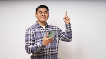 Photo for Young Asian man using smartphone over white background with a big smile on face, pointing with hand finger to the side looking at the camera. studio shot - Royalty Free Image