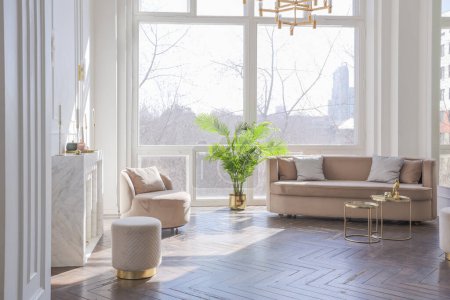 sehr helles und helles Interieur eines luxuriösen gemütlichen Wohnzimmers mit schicken weichen beigen Möbeln mit goldenen Metallic-Elementen, riesigem Fenster zum Boden und Holzparkett
