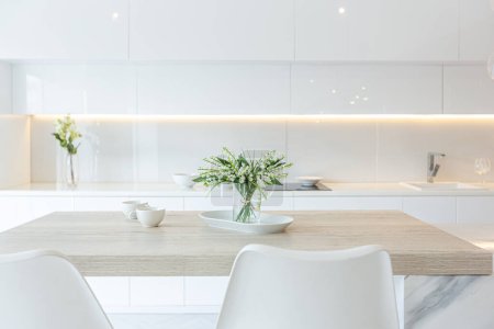 Luxus-Interieur der modernen trendigen schneeweißen Küche im minimalistischen Stil mit Insel und zwei Barhockern. Riesige Fenster zum Boden und ein Glasregal für Geschirr