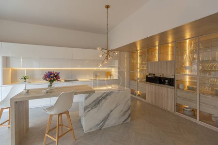 Luxus-Interieur der modernen trendigen schneeweißen Küche im minimalistischen Stil mit Insel und zwei Barhockern. Riesige Fenster zum Boden und ein Glasregal für Geschirr