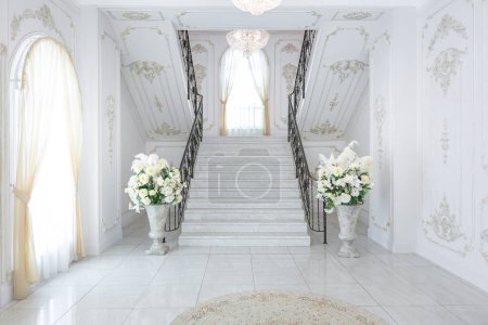 Téléchargez les photos : Luxueux intérieur royal chic dans un style baroque. hall très lumineux, lumineux et blanc avec meubles de style ancien coûteux. escalier en marbre large chic menant au deuxième étage - en image libre de droit