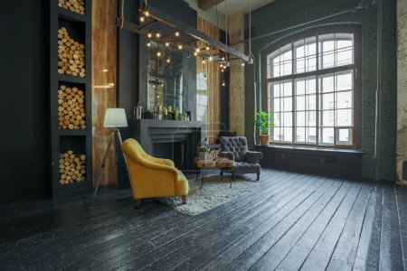 Foto de Oscuro interior brutal de la sala de estar decorada con troncos de madera. sillones suaves amarillos y grises, gran ventana de arco y chimenea - Imagen libre de derechos