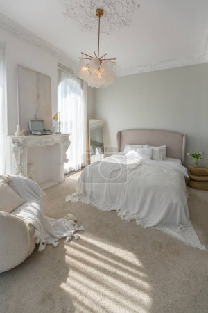 Foto de Un apartamento acogedor y luminoso con enormes ventanas panorámicas con luz solar. dormitorio en tonos beige suaves con chimenea y un cómodo y elegante sillón tapizado y cama - Imagen libre de derechos