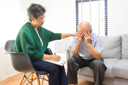 Foto de Hombre mayor asiático deprimido compartiendo problemas durante la sesión de terapia con psiquiatra mujer escribiendo en portapapeles. - Imagen libre de derechos