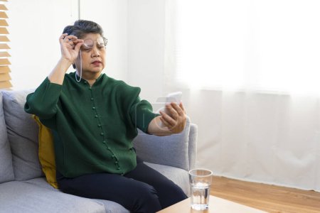 Téléchargez les photos : Femme âgée ayant des problèmes avec la vue longue en essayant de lire un livre, assis sur le canapé à la maison. - en image libre de droit