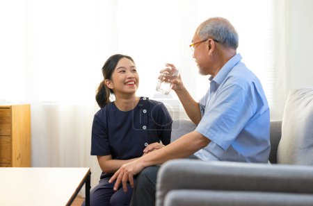 Téléchargez les photos : Infirmière souriante donnant un verre d'eau à un homme asiatique âgé dans une maison de soins infirmiers ou un centre d'aide à la vie autonome. - en image libre de droit
