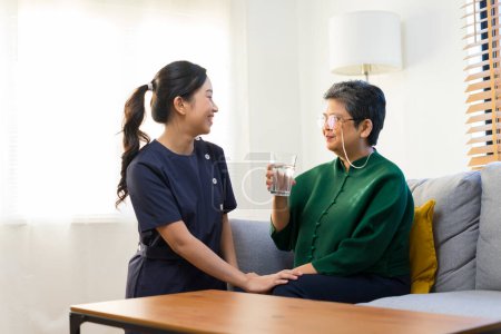 Photo for Smiling nurse giving glass of water to senior asian woman in nursing home or assisted living facility - Royalty Free Image