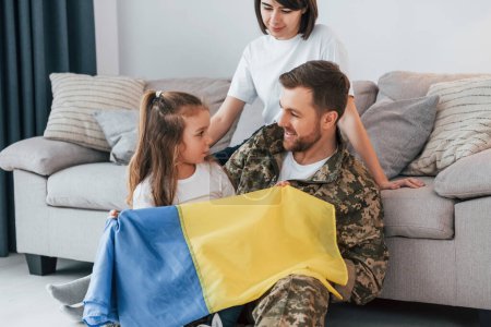 Photo for With Ukrainian flag. Soldier in uniform is at home with his wife and daughter. - Royalty Free Image