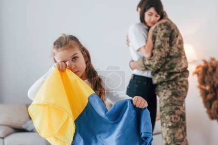 Téléchargez les photos : Fille debout devant son parent et tenant le drapeau de l'Ukraine. Soldat en uniforme est à la maison avec sa femme et sa fille. - en image libre de droit