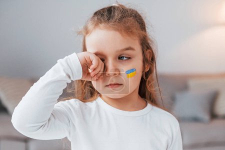 Foto de Un niño inocente está llorando. Retrato de niña con bandera ucraniana maquillaje en la cara. - Imagen libre de derechos