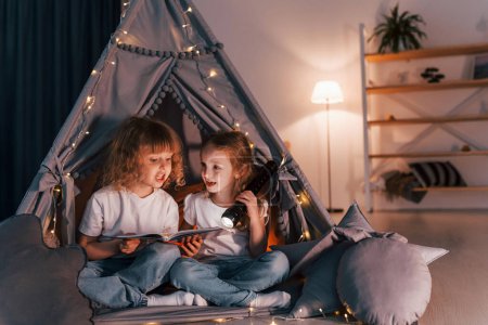 Une fille tenant une lampe torche, l'autre lisant le livre. Deux petits amis est dans la tente dans la chambre domestique ensemble.