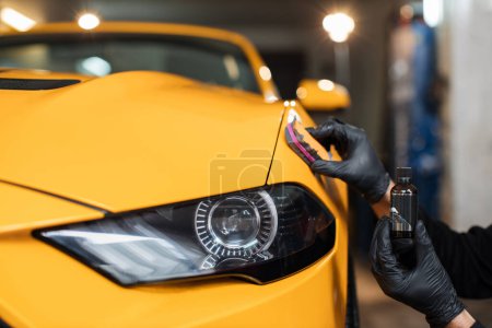 Photo for Cropped close up image of male hand in protective black glove, holding sponge with solid carnauba wax, and polishing hood of luxury yellow car at professional detailing workshop. Car detailing. - Royalty Free Image