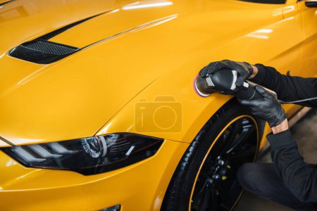 Auto detailing service, polishing of the car. Close up view of hands of man worker, polishing hood and body of modern yellow luxury car with orbital polisher and wax.