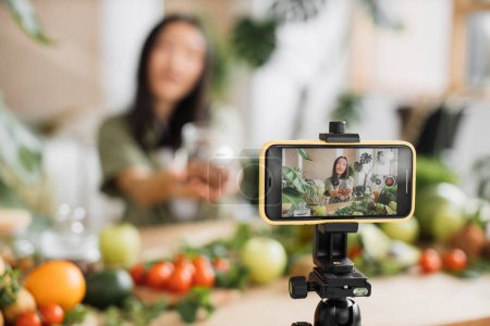 Photo for Selective focus on smartphone, young asian woman blogger or content creator preparing healthy salad and recording video on phone. Attractive female holding hazelnuts while recording blog. - Royalty Free Image