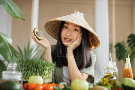 Foto de Mujer asiática alegre en tradicional sombrero cónico haciendo ensalada de verduras y frutas disfrutando de una cena saludable con aguacate en exótico estudio de luz en un resort tropical. Alimentación y nutrición para bajar de peso. - Imagen libre de derechos