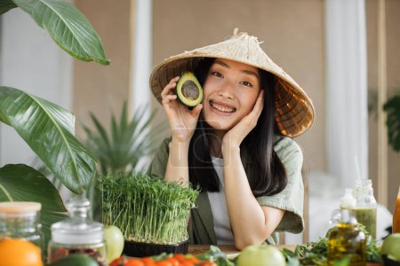 Foto de Mujer asiática alegre en tradicional sombrero cónico haciendo ensalada de verduras y frutas disfrutando de una cena saludable con aguacate en exótico estudio de luz en un resort tropical. Alimentación y nutrición para bajar de peso. - Imagen libre de derechos