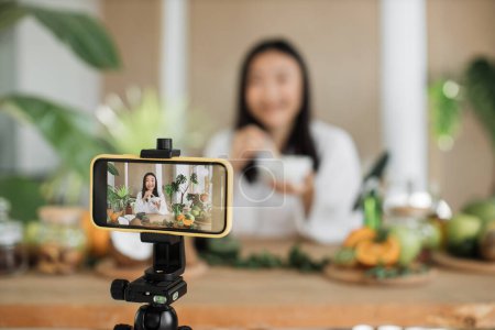 Photo for Young asian woman blogger or content creator in white bathrobe sitting at wooden table with various ingredients preparing natural cosmetics at home, mixing the mass in a bowl while preparing cream. - Royalty Free Image