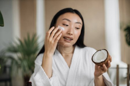 Foto de Vista de cerca de la cara de la joven mujer asiática que aplica cosméticos naturales en casa, la celebración de coco para la crema casera para la piel, el cuidado del cabello con un fondo exótico en el balneario - Imagen libre de derechos