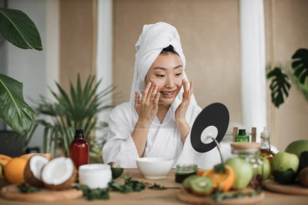 Foto de Mujer asiática saludable en albornoz blanco y toalla sentada en la mesa de madera con varios ingredientes que preparan cosméticos naturales en casa mezclando masa en un tazón mientras preparan crema y aplican en la cara. - Imagen libre de derechos