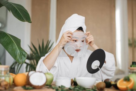 Foto de Atractiva mujer asiática mirando el espejo sentado en la mesa con ingredientes para cosméticos caseros aplicando mascarilla de algodón en su cara. Jovencita haciendo procedimientos anti envejecimiento. - Imagen libre de derechos