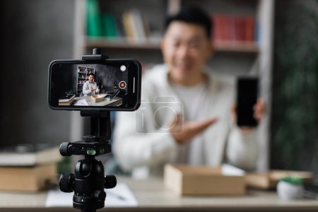 Téléchargez les photos : Focus sur l'écran du smartphone, homme asiatique agréable filmer la vidéo sur la caméra de téléphone moderne tout en ouvrant boîte de colis avec nouveau smartphone. Concept de personnes, de technologie et de blogging. - en image libre de droit