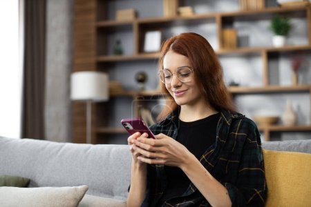 Photo for Portrait of young caucasian woman sitting on cozy sofa with modern smartphone in hands. Pretty female resting during working day from home. - Royalty Free Image