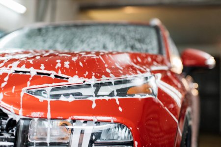 Photo for Washing a car headlight at auto wash service. Soapy red car. Washing of crossover car with active cleaning foam, front view of car soapy hood. - Royalty Free Image