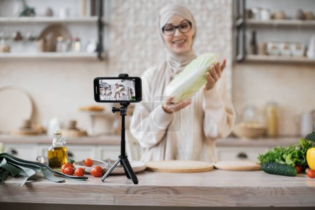 Téléchargez les photos : Concentrez-vous sur l'écran. Jeune blogueuse musulmane tenant du chou pékin, streaming en direct et enregistrement vidéo recette pour blog alimentaire. - en image libre de droit