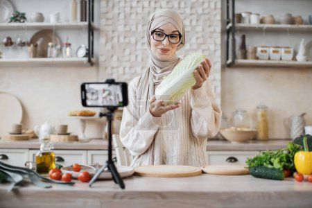 Téléchargez les photos : Jeune blogueuse musulmane tenant du chou pékin, streaming en direct et enregistrement vidéo recette pour blog alimentaire. Jeune jolie femelle dans hijab cuisson des aliments en ligne à la maison cuisine. - en image libre de droit