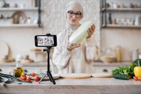 Téléchargez les photos : Concentrez-vous sur l'écran. Jeune blogueuse musulmane tenant du chou pékin, streaming en direct et enregistrement vidéo recette pour blog alimentaire. - en image libre de droit
