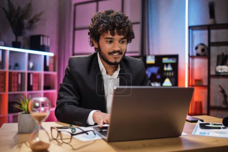 Téléchargez les photos : Handsome experienced smart stylish curly bearded manager, broker, businessman, wearing suit sitting at table, using laptop at evening time in modern office. - en image libre de droit