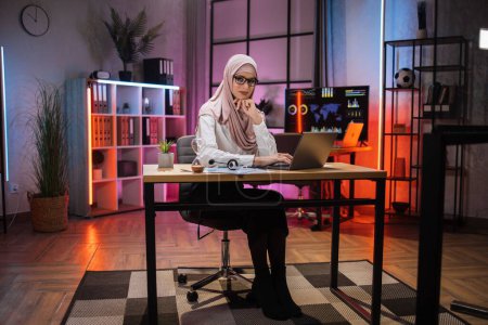 Photo for People, overwork, deadline and office work concept. Confident smiling muslim woman in beige hijab sitting at desk with modern laptop and looking at camera at evening time at office. - Royalty Free Image