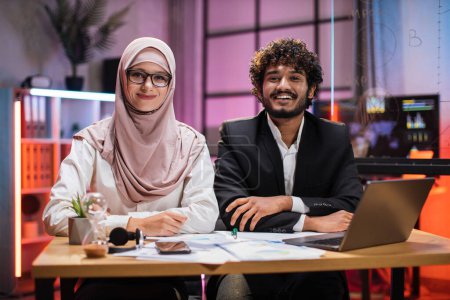 Foto de Confident two multi ethnic economists, young experienced muslim female and businessman, office manager in formal wear sitting at table using laptop in evening office. - Imagen libre de derechos
