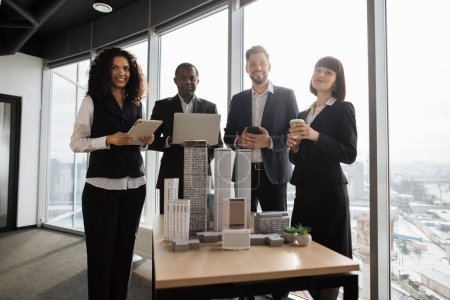 Foto de Team of diverse four multiethnic businesspeople having meeting in boardroom at office with panoramic windows blurry cityscape standing near table with skyscraper building maquette looking at camera - Imagen libre de derechos