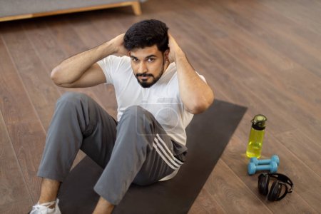 Téléchargez les photos : Homme indien attrayant vêtu de vêtements de sport en utilisant un tapis de yoga pour faire des sit-ups dans le salon. Entraînement à domicile pour les muscles abdominaux. Auto-discipline et concept de motivation. - en image libre de droit
