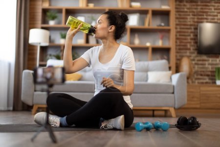 Photo for Focused african american female in gym clothes drinking water from sports bottle while sitting cross-legged in front of active cell phone on tripod. Fit sports vlogger showing workout routine at home. - Royalty Free Image