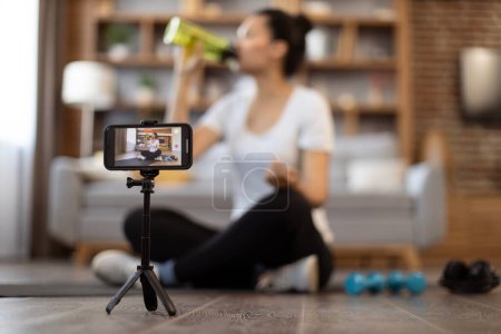 Photo for Modern smartphone on tripod recording video of young multiracial lady in athletic wear drinking from sports bottle on camera. Media influencer shooting video for her sports vlog followers indoors. - Royalty Free Image