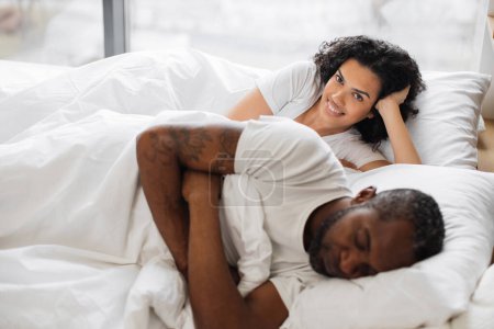 Photo for Pretty african american woman leaning on soft pillow already awake while her attractive husband still getting shuteye on bed in front of panoramic window. Happy couple spending morning together. - Royalty Free Image