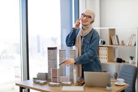 Photo for Happy muslim adult wearing hijab and casual wear speaking on mobile while reaching scale model of apartment building with pencil in hand. Female designer consulting about residential project plan. - Royalty Free Image