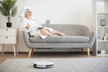 Foto de Mujer sonriente joven en albornoz blanco tumbado en el sofá con el ordenador portátil y el robot de funcionamiento aspiradora sobre el control remoto que muestra el pulgar hacia arriba. - Imagen libre de derechos
