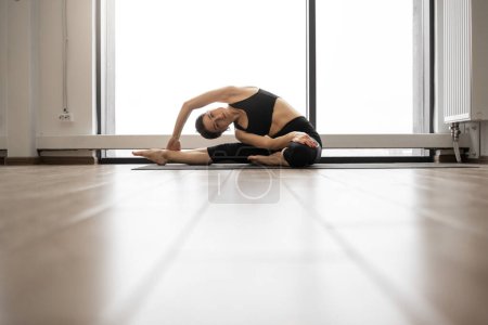 Kaukasische schlanke Frau, die die Seiten zu ausgestreckten Beinen beugt, während sie auf einer Yogamatte in einem geräumigen Raum sitzt. Fitness junge Dame in schwarzer Sportbekleidung macht Übungen zur Dehnung des Körpers.