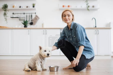 Photo for Portrait of beautiful relaxed woman and well-trained white terrier posing in modern kitchen of spacious house. Confident pet owner finishing brief indoor training session with homemade treats. - Royalty Free Image