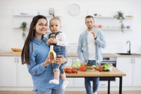Photo for Precious little cutie with ponytail eating organic banana while sitting on mommys hands. Responsible father cutting and cooking vegetables for lunch on wooden table in white kitchen. - Royalty Free Image