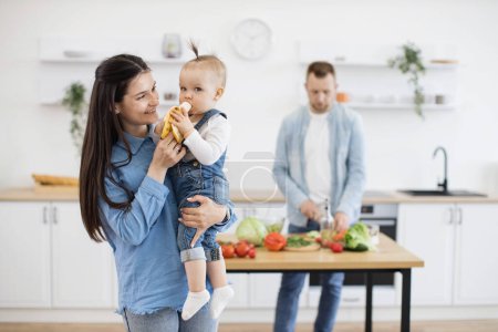 Photo for Precious little cutie with ponytail eating organic banana while sitting on mommys hands. Responsible father cutting and cooking vegetables for lunch on wooden table in white kitchen. - Royalty Free Image