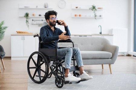 Photo for Full length view of adult person in wheelchair talking on cell phone while using comfort of modern studio flat. Mature arabian man organising birthday party while getting recovery from illness. - Royalty Free Image