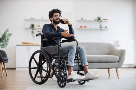Photo for Full length view of adult person in wheelchair talking on cell phone while using comfort of modern studio flat. Mature arabian man organising birthday party while getting recovery from illness. - Royalty Free Image