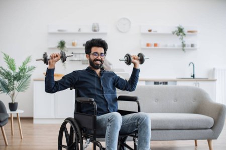Photo for Joyous bearded man in wheelchair exercising with weights while following workout program at home. Positive indian adult in glasses and cozy outfit participating in physical activity in apartment. - Royalty Free Image
