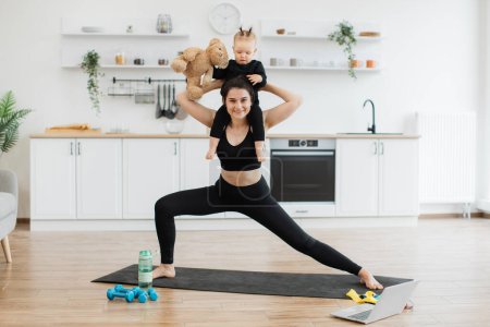 Photo for Full length portrait of joyous lady posing with infant sitting around neck with teddy bear in dining room. Healthy strong mother achieving yoga pose with double weights of daughter and soft companion. - Royalty Free Image